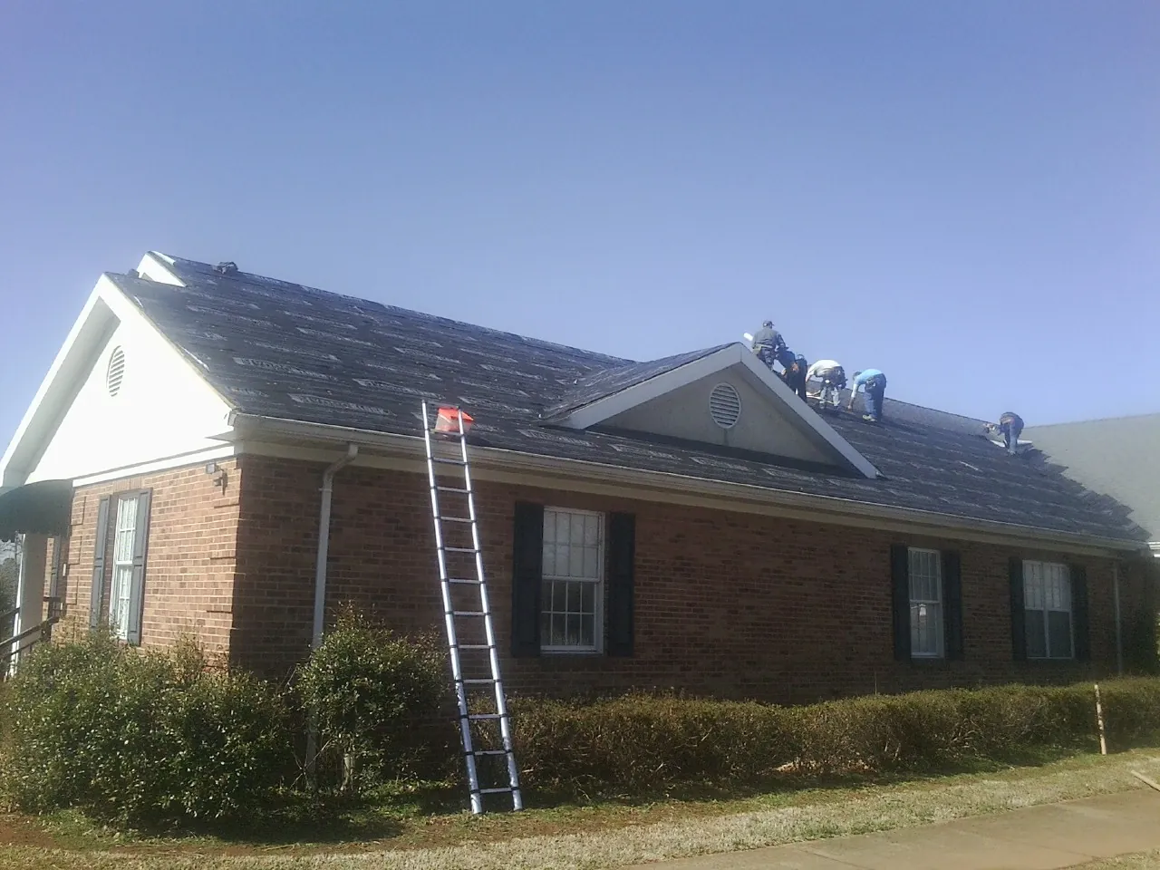 Crew tearing shingles off a brick ranch roof with a ladder set — Metal Roof Repair work by our crew in Algonac