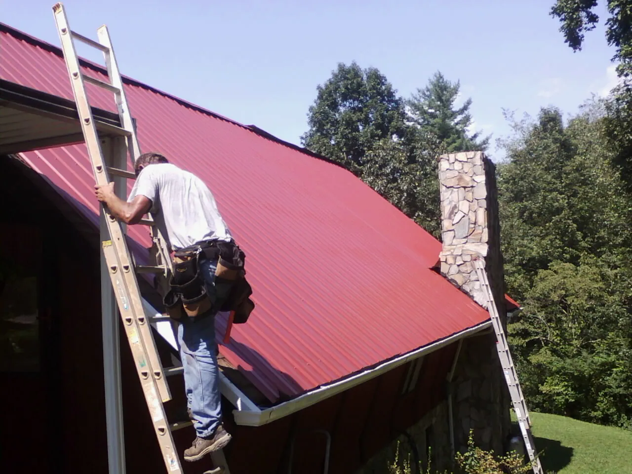 Roofer on a ladder beside a red metal roof and stone chimney — Soffit & Fascia Repair by Roofing Brothers in Algonac